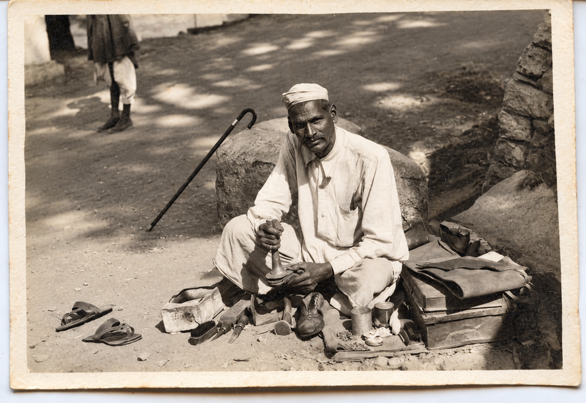 A shoemaker sitting on the ground outdoors, working on a shoe with tools and materials spread before him.