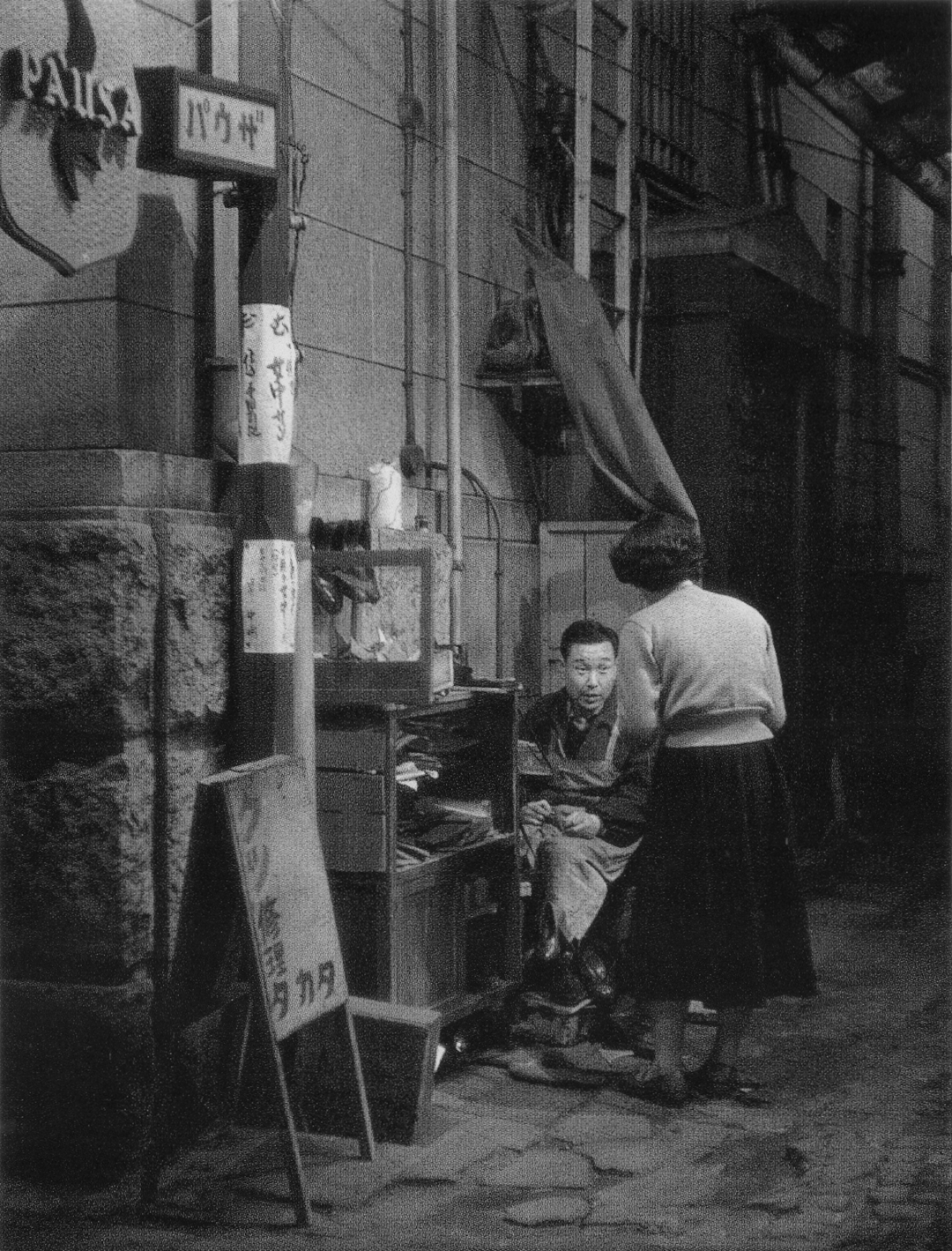 A woman stands in a narrow alleyway speaking to a man sitting in a small, street-side repair booth.