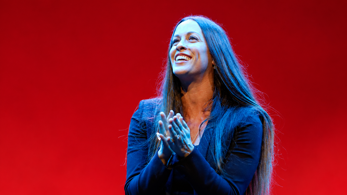 Alanis Morissette smiling and clapping in front of a red background.