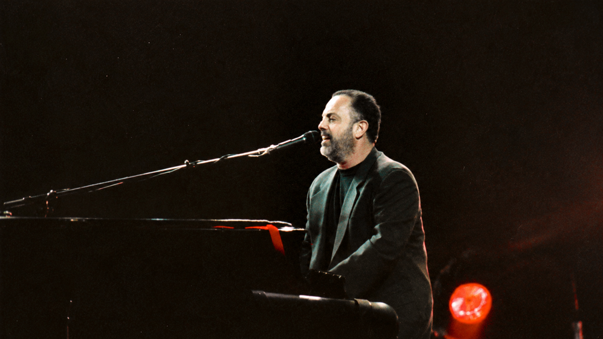 Celebrities betrayed my managers. Billy Joel singing into a microphone while playing the piano on a dark stage.