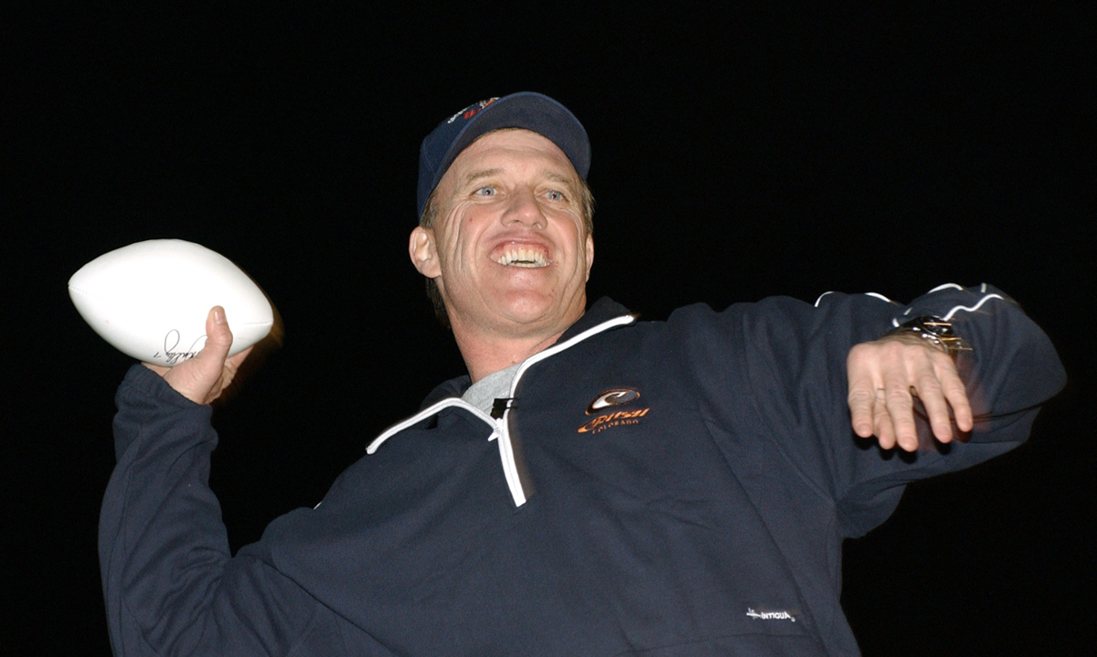 John Elway wearing a blue Colorado Rockies quarter-zip jacket and baseball cap, smiling while holding an autographed football.