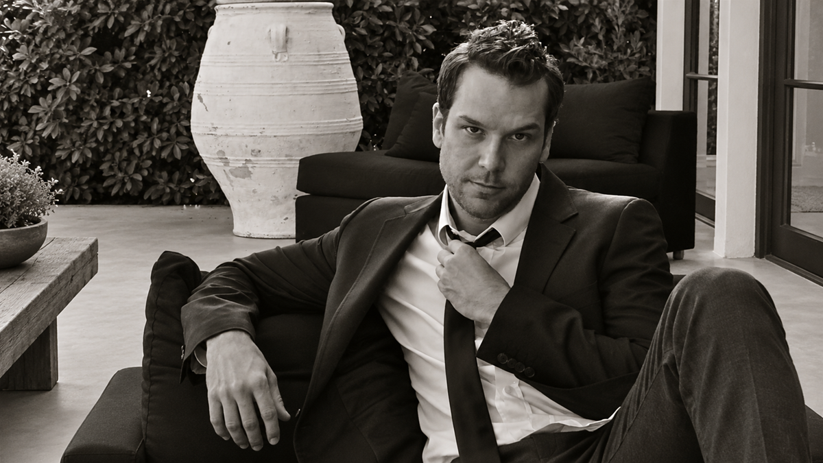 A man in a suit and white shirt sits on an outdoor sofa, adjusting his tie while looking at the camera.
