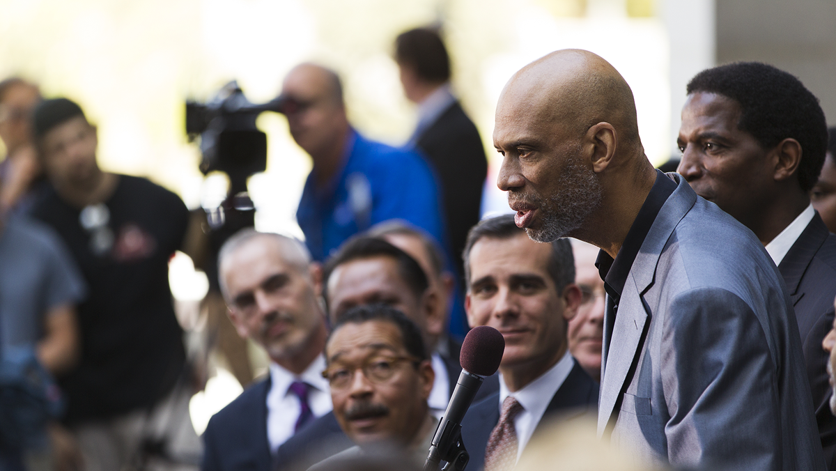 Kareem Abdul-Jabbar speaks into a microphone at a press conference surrounded by a crowd of people.
