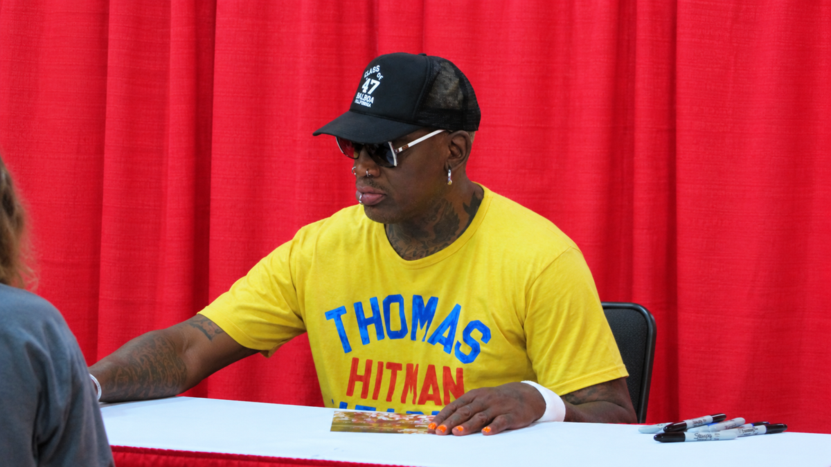 Dennis Rodman wearing a yellow t-shirt and a black baseball cap sits at a table to sign an autograph.