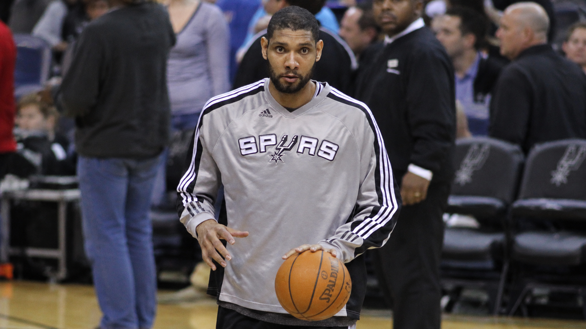 Tim Duncan in a grey San Antonio Spurs warm-up shirt dribbles a basketball on an indoor court.