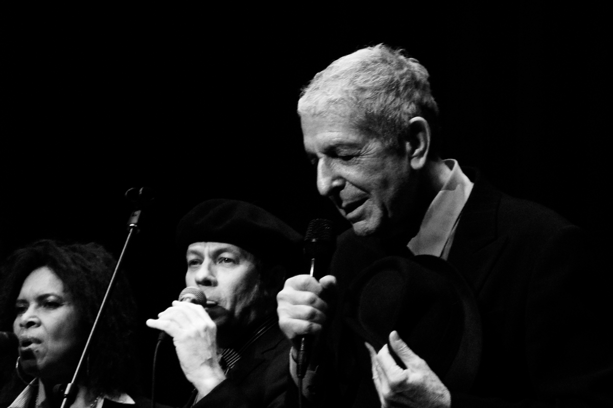 Leonard Cohen sings into a microphone on stage, accompanied by two backup vocalists.