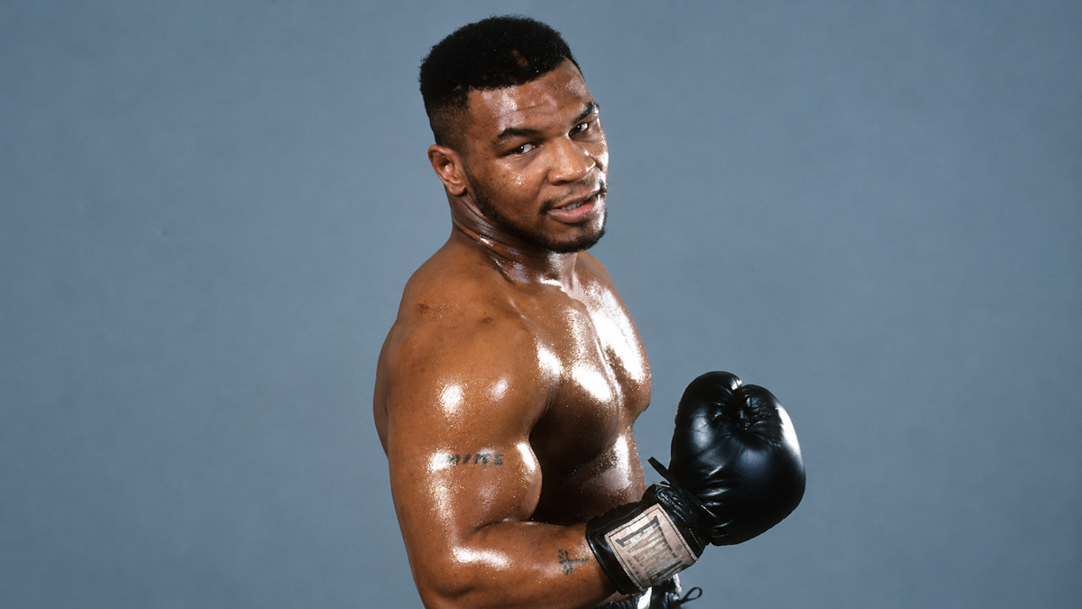 A shirtless, muscular Mike Tyson wearing black boxing gloves and looking toward the camera.