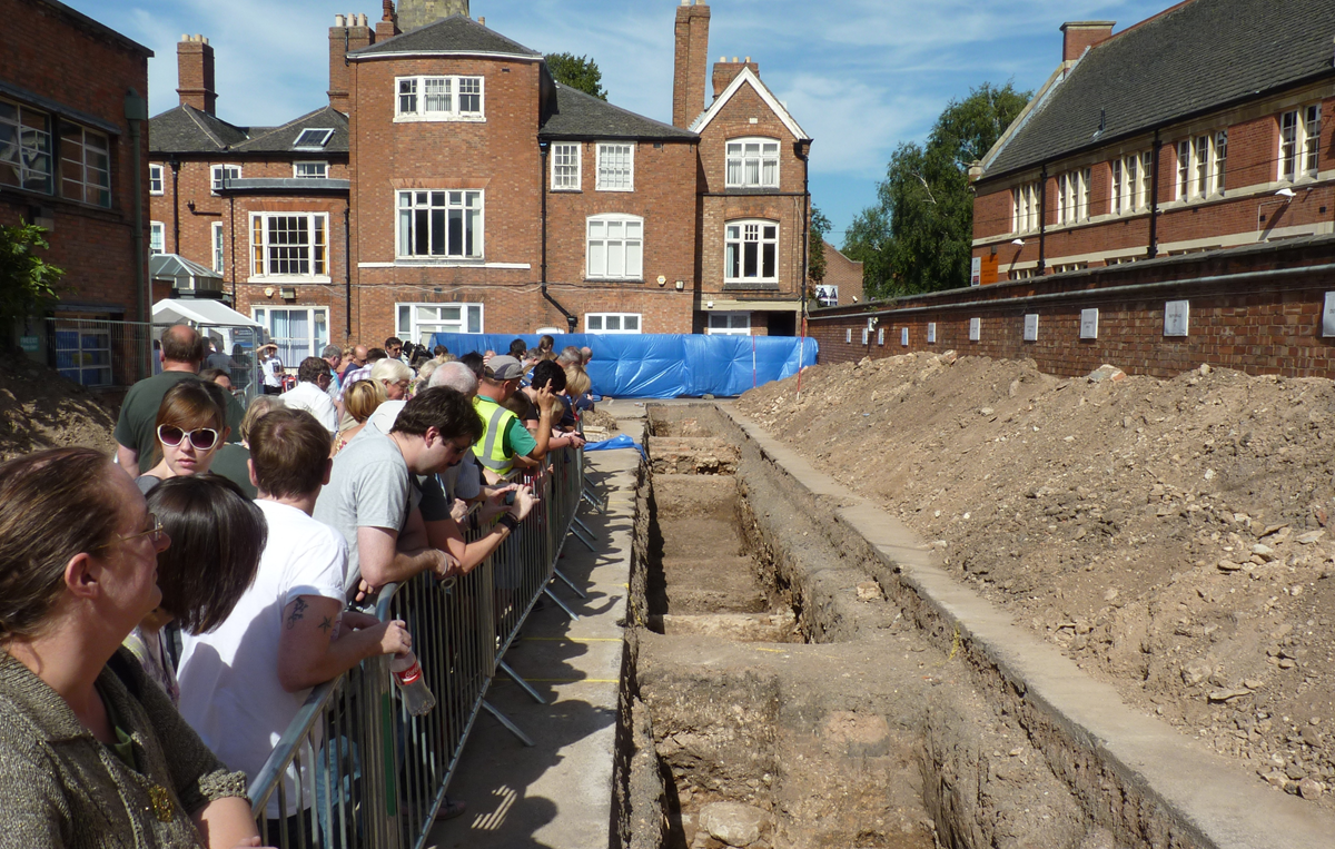 A crowd of people stands behind a metal barrier, watching an archaeological excavation trench in front of a brick building.