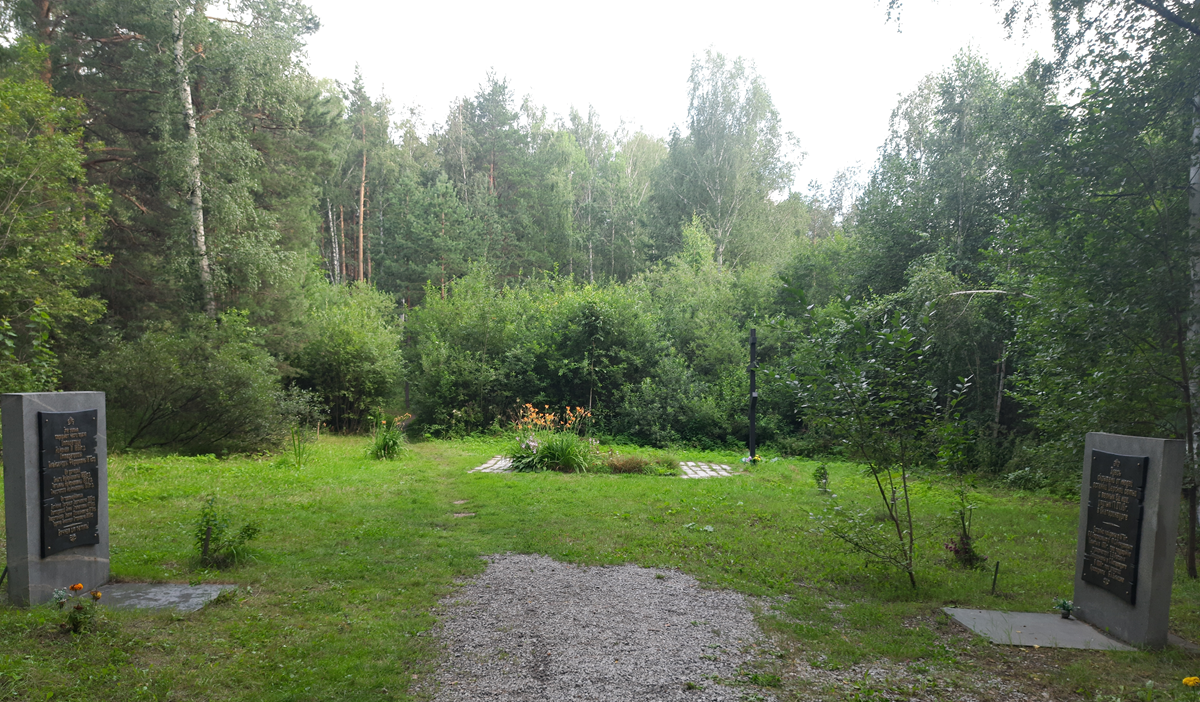 Two stone monuments with memorial plaques stand on either side of a gravel path leading into a clearing surrounded by trees.