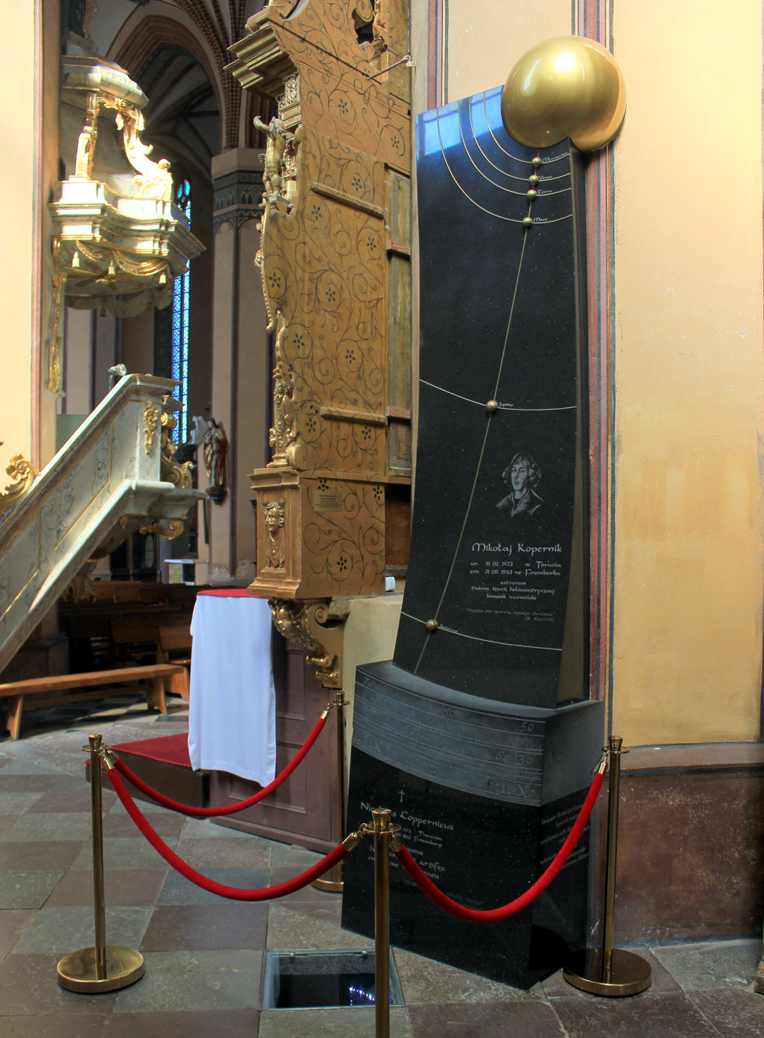 A black stone monument dedicated to Nicolaus Copernicus, featuring an engraved solar system diagram, a portrait, and text, standing in a church near a decorative wooden structure.