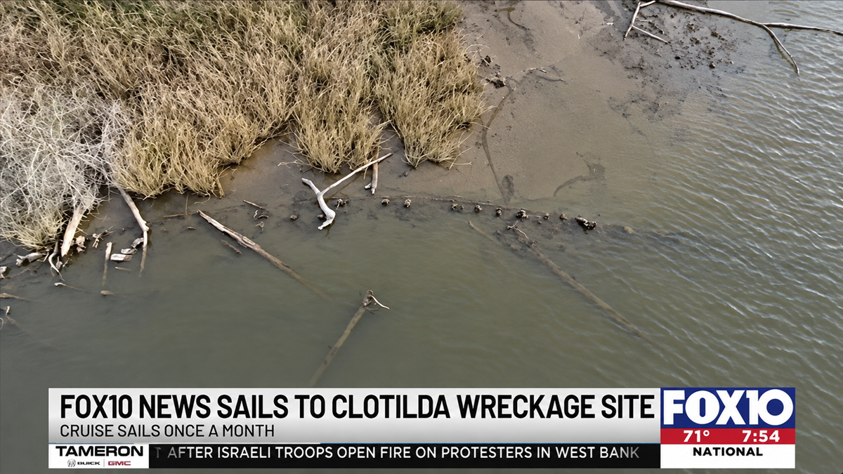 An aerial view of the muddy shoreline where the Clotilda shipwreck remains are partially submerged in the water, with a Fox 10 news graphic at the bottom of the screen.