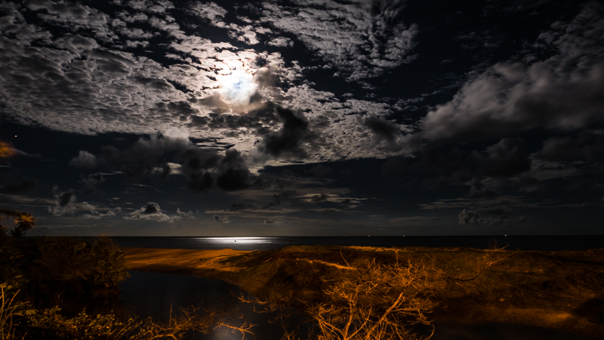 Full moon shining through cloudy night sky above a dark beach and ocean.
