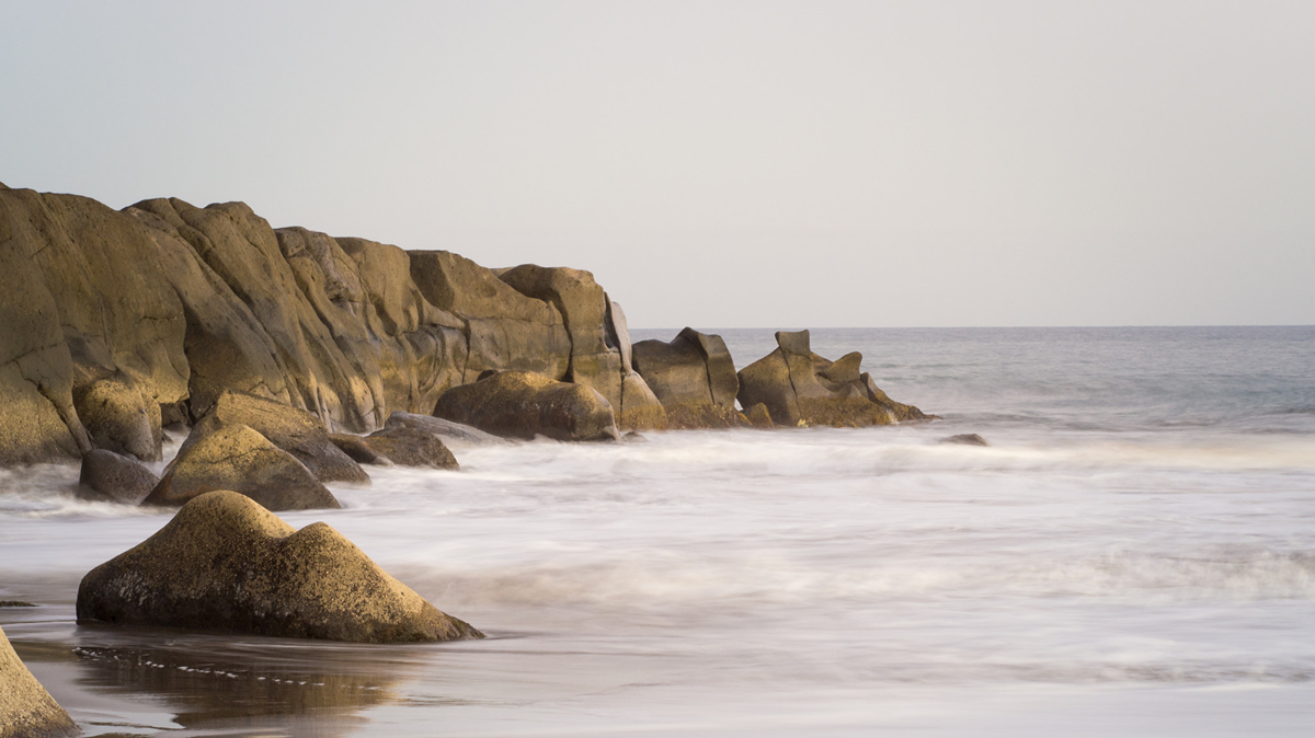 Large rocks along a beach with waves washing over the sand.