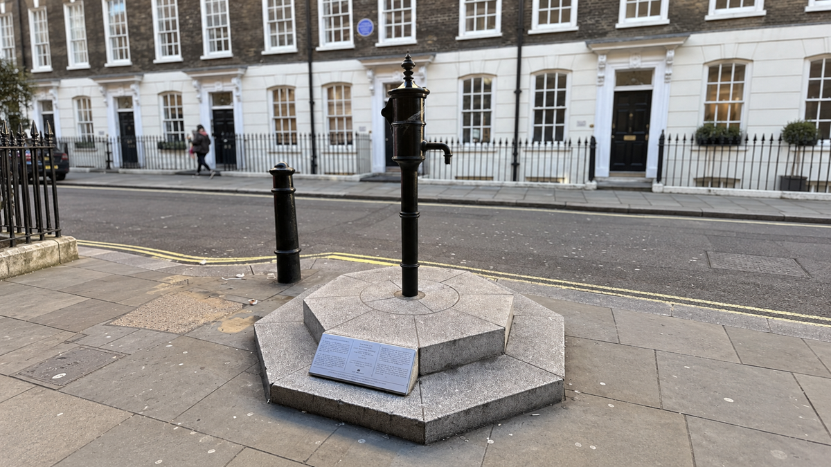 A historic black cast-iron water pump standing on a stone base on a London sidewalk, with a commemorative plaque attached to the front.