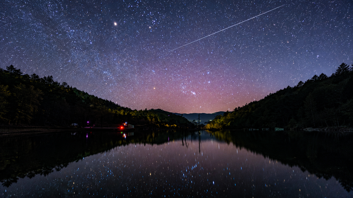 Starry night sky with a long streak of light, likely a meteor or satellite, reflecting in a calm lake between forested hills.