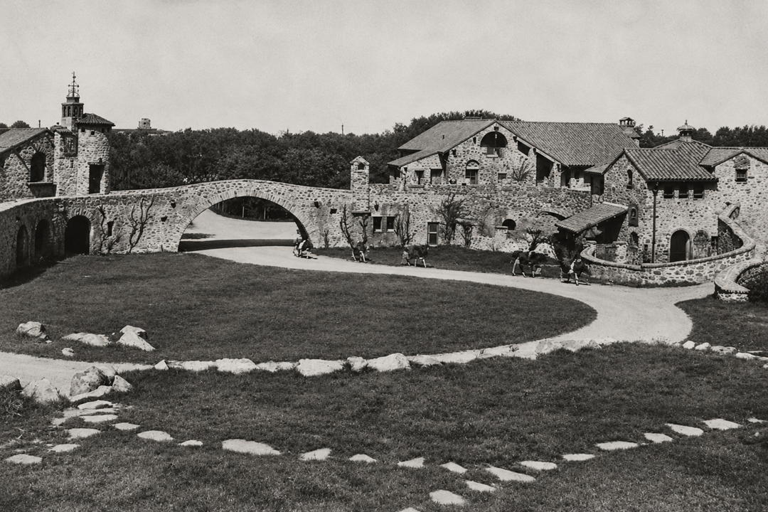 A stone house with an arched entryway and courtyard, featuring a circular driveway and scattered groups of people walking on the grounds.