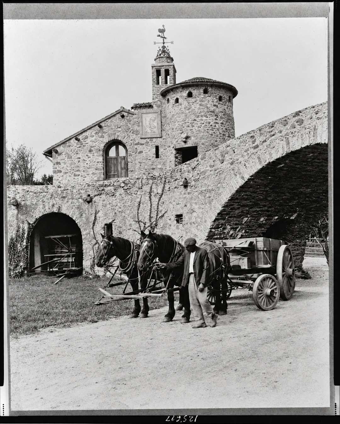 A man stands beside a team of two horses harnessed to a wooden wagon in front of a stone building with an arched bridge.
