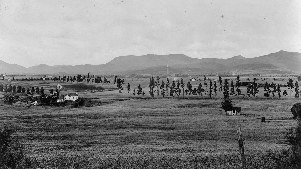 Black and white panoramic view of a wide valley with scattered trees, farm buildings, and a windmill, framed by distant mountains.