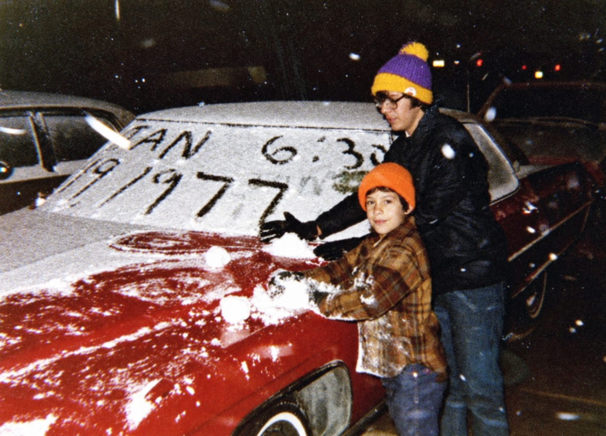 Two people in winter hats make snowballs on the hood of a red car, which has the date “JAN 6:30 1977” written in the frost on the windshield.