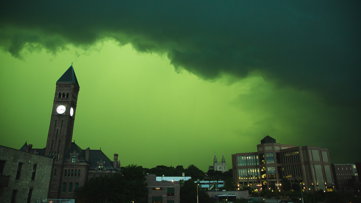 A cityscape with a tall clock tower and large buildings under a dramatic, vibrant green stormy sky.