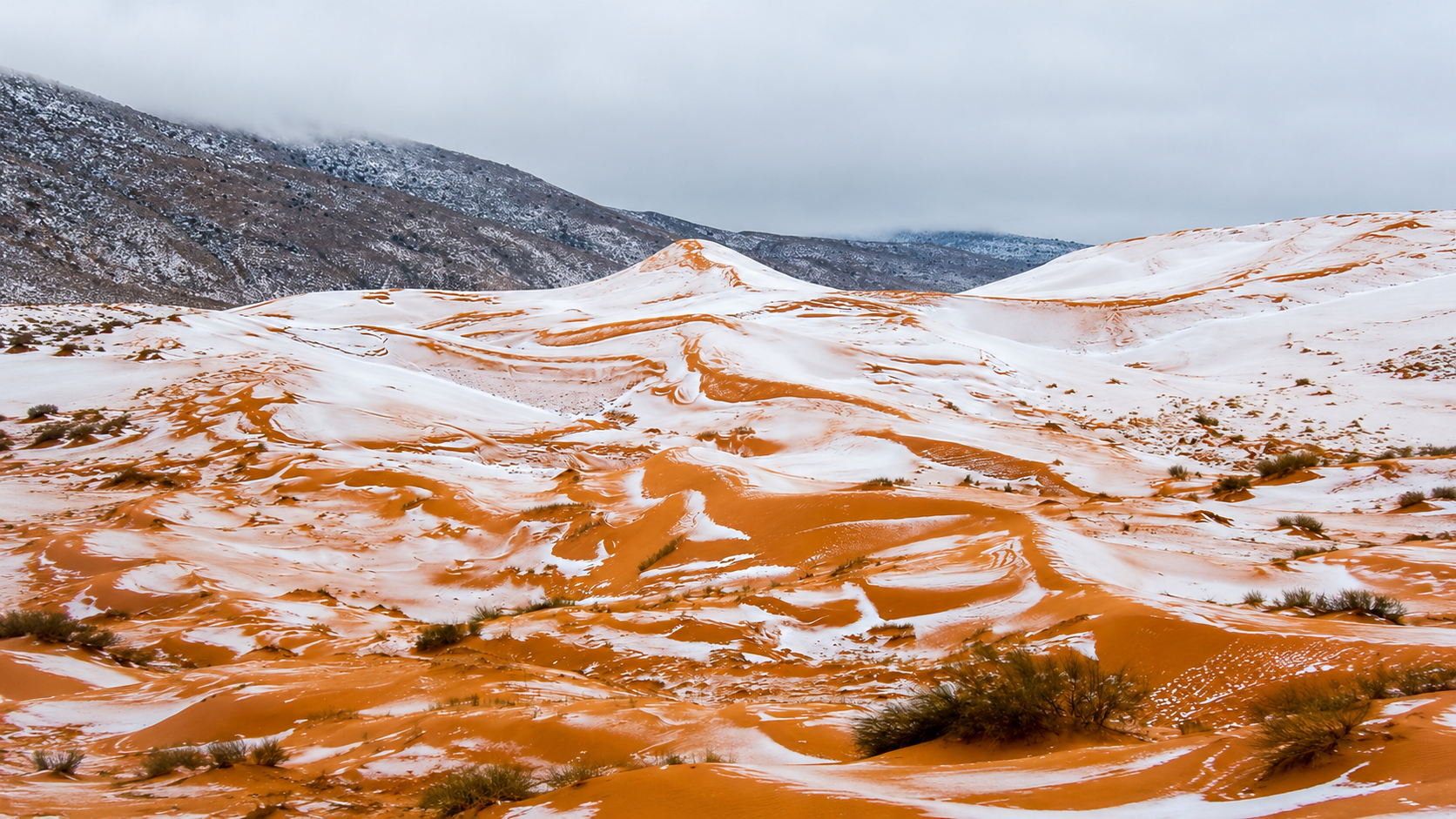 Snow-dusted orange sand dunes beneath a cloudy sky and mountains.
