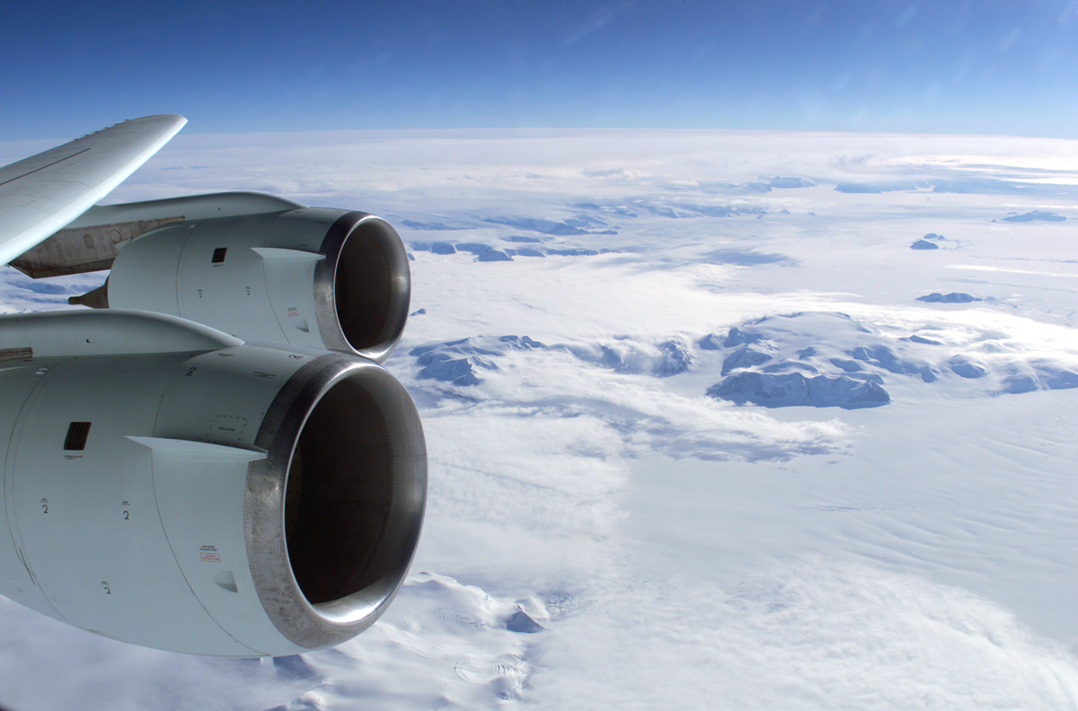Two jet engines mounted under an airplane wing, seen from above snowy mountains and clouds.