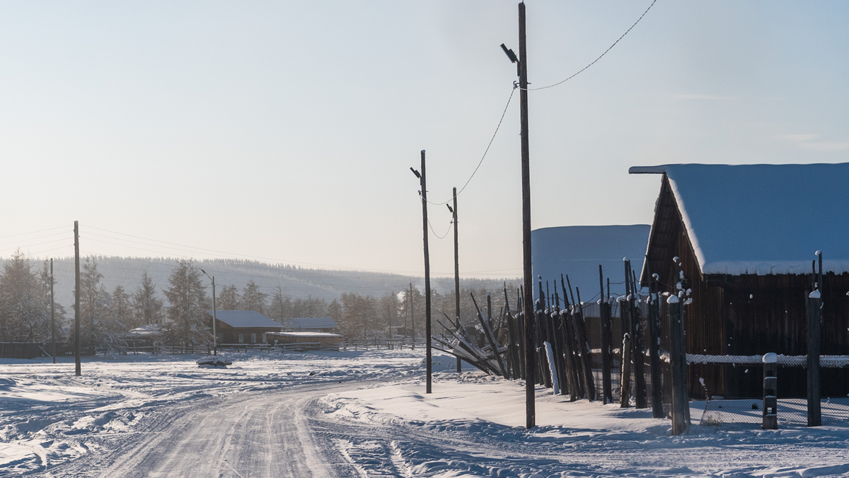 Coldest cities. A snow-covered road leads past wooden houses and utility poles in a rural landscape during winter.