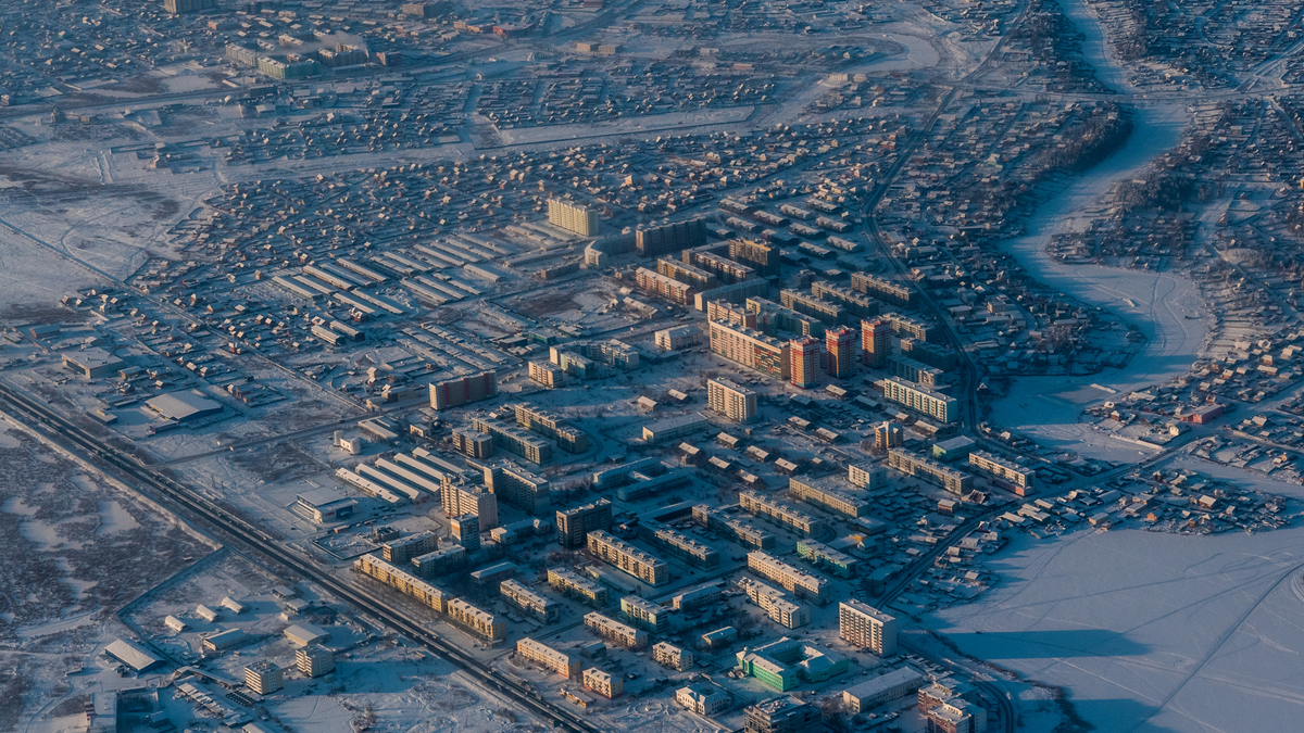 Aerial view of a town covered in snow, featuring residential neighborhoods, apartment blocks, and a frozen river winding through the landscape.