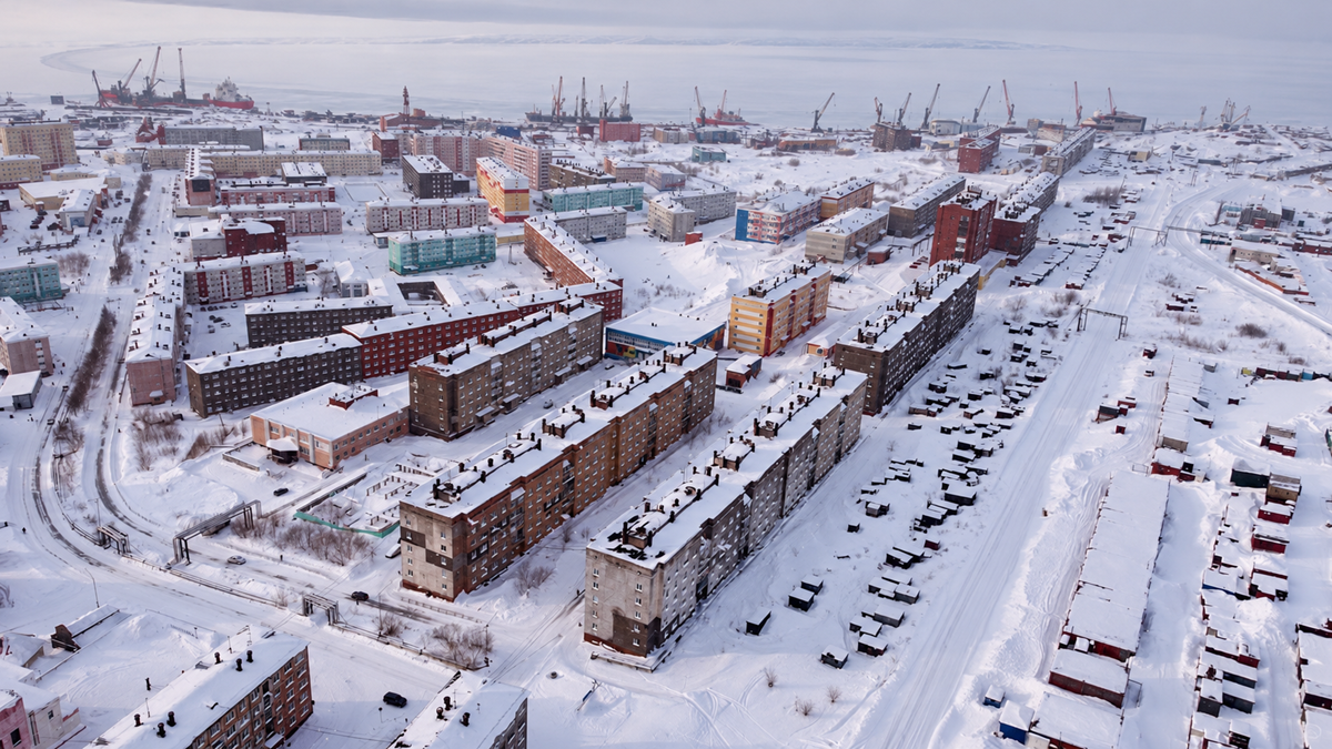 Aerial view of an arctic town with block apartment buildings and roads covered in deep snow, with a harbor and cranes in the background.