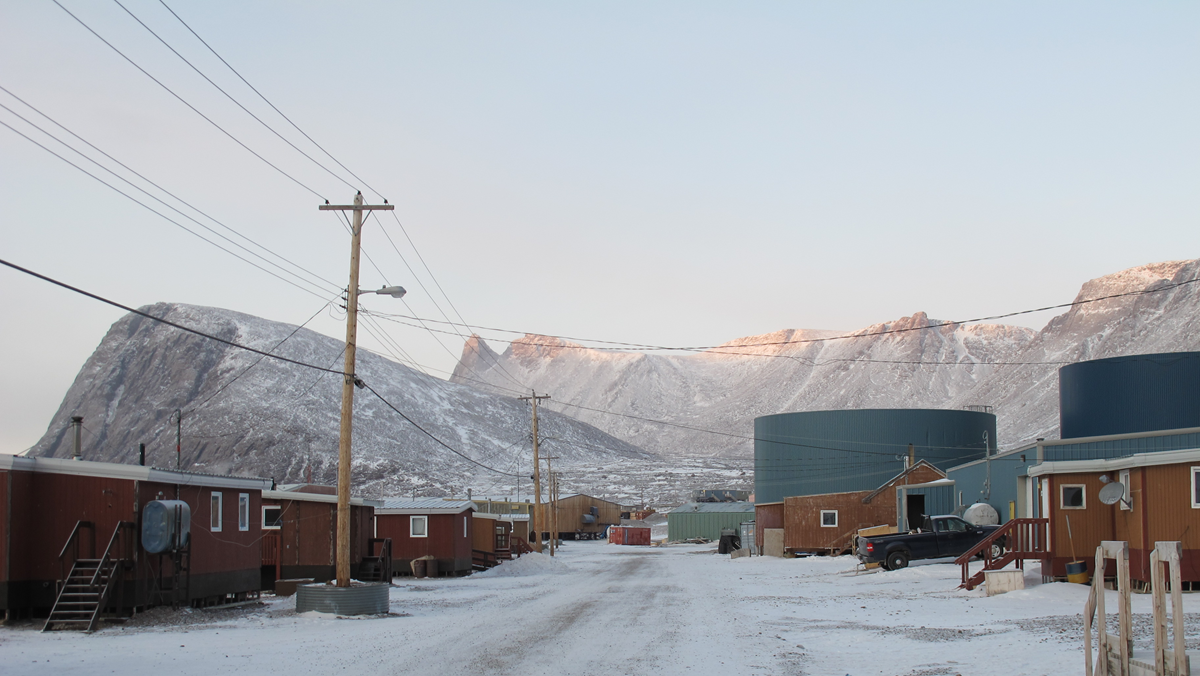 A snowy street in a remote town flanked by small, prefabricated buildings, large storage tanks, and high, snow-covered mountains.