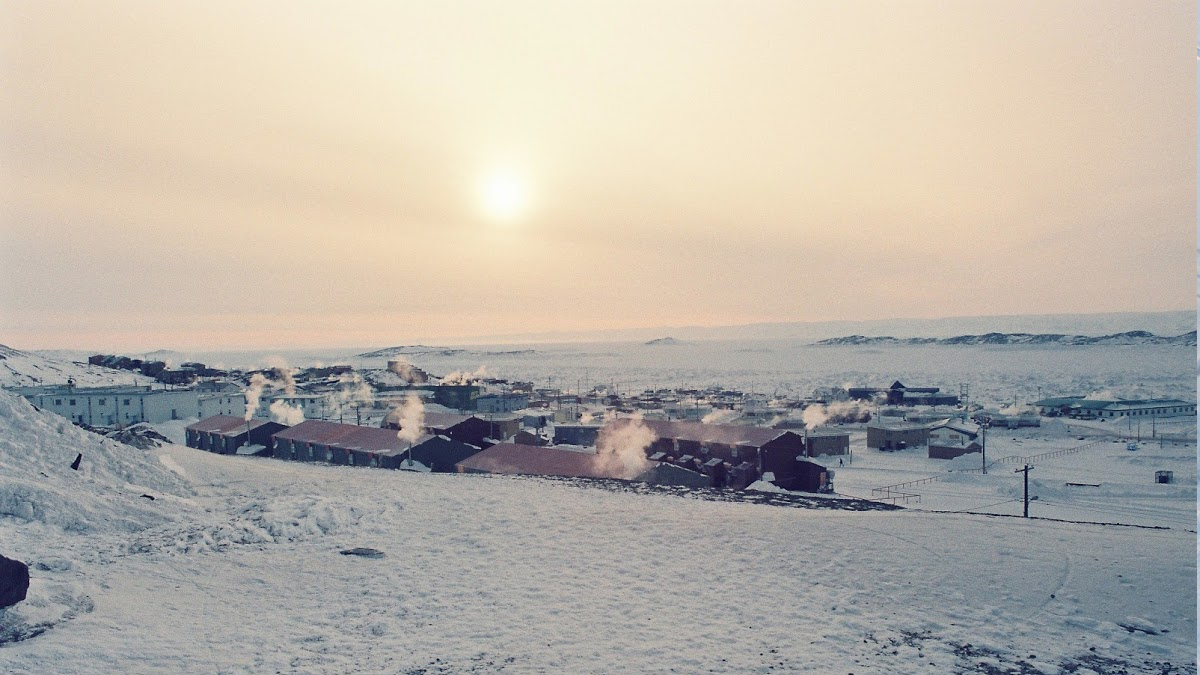 A remote town with snow-covered buildings and steaming chimneys under a pale sun.