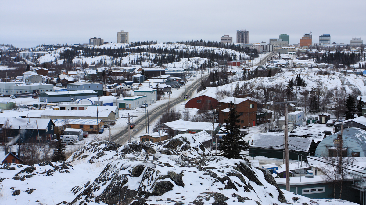 A high-angle view of a town in winter, with snow-covered rocks in the foreground, roads winding through buildings, and a skyline of buildings in the distance.