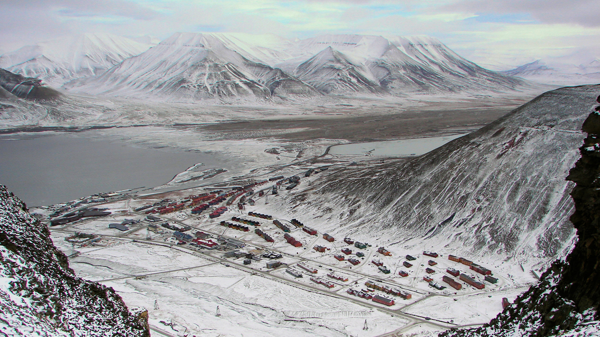 A high-angle view of Longyearbyen, a town with colorful buildings nestled in a snowy valley beneath large, snow-covered mountains.