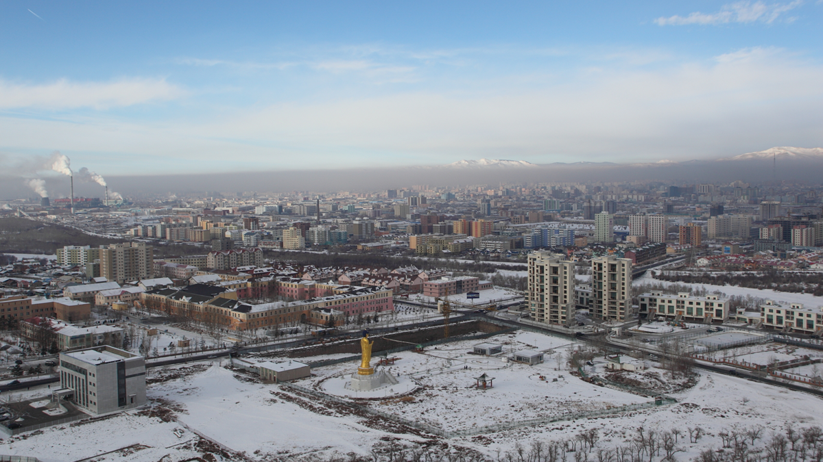 An elevated view of a snow-covered city featuring a prominent golden Buddha statue in the foreground and a hazy skyline with mountains in the distance.