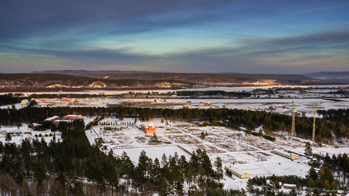 Aerial view of a snow-covered landscape with scattered buildings, a dense pine forest, and a distant mountain range under a dusk sky.