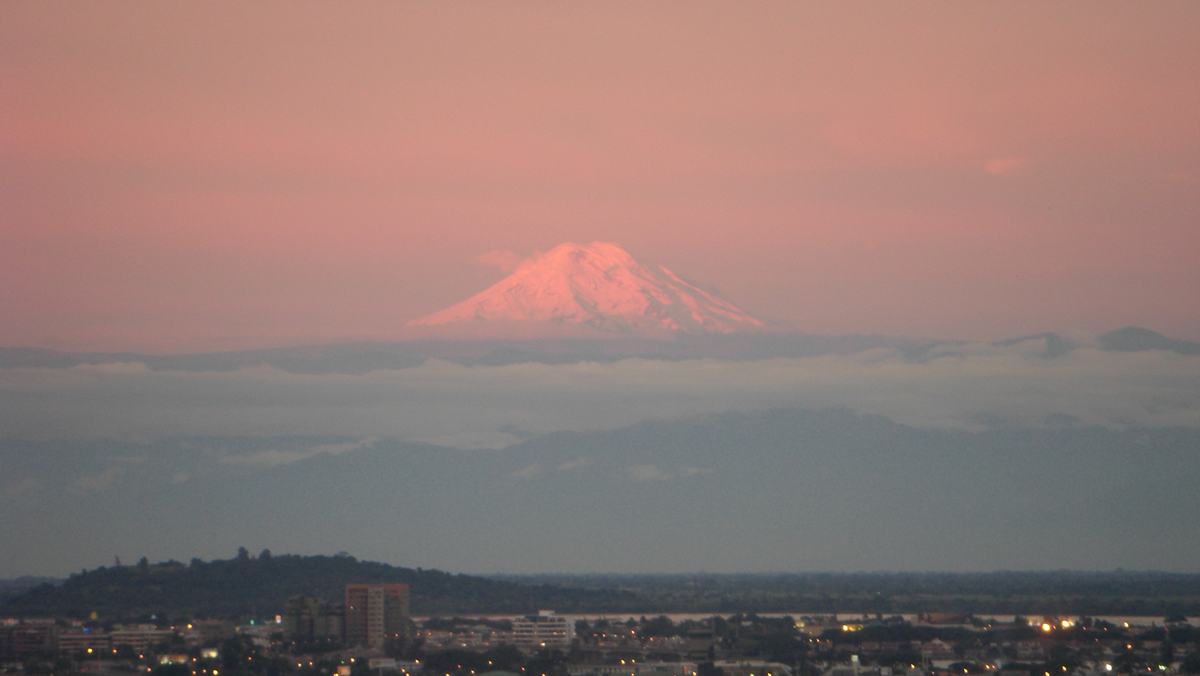 A snow-capped volcano peak rising above a layer of clouds at sunset, with a city visible in the foreground.