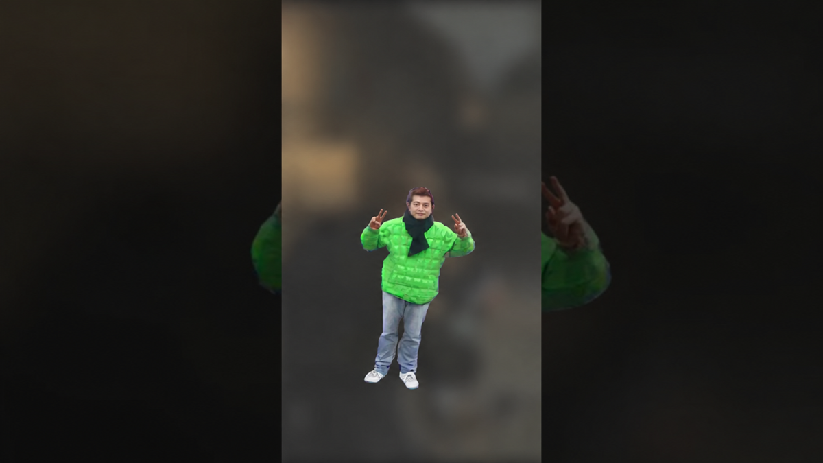 A person in a bright green puffer jacket, black scarf, and jeans holds up peace signs with both hands.