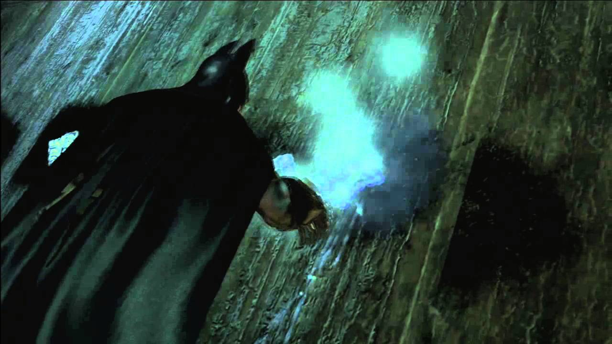 Batman standing against a wall while using a glowing blue gadget.
