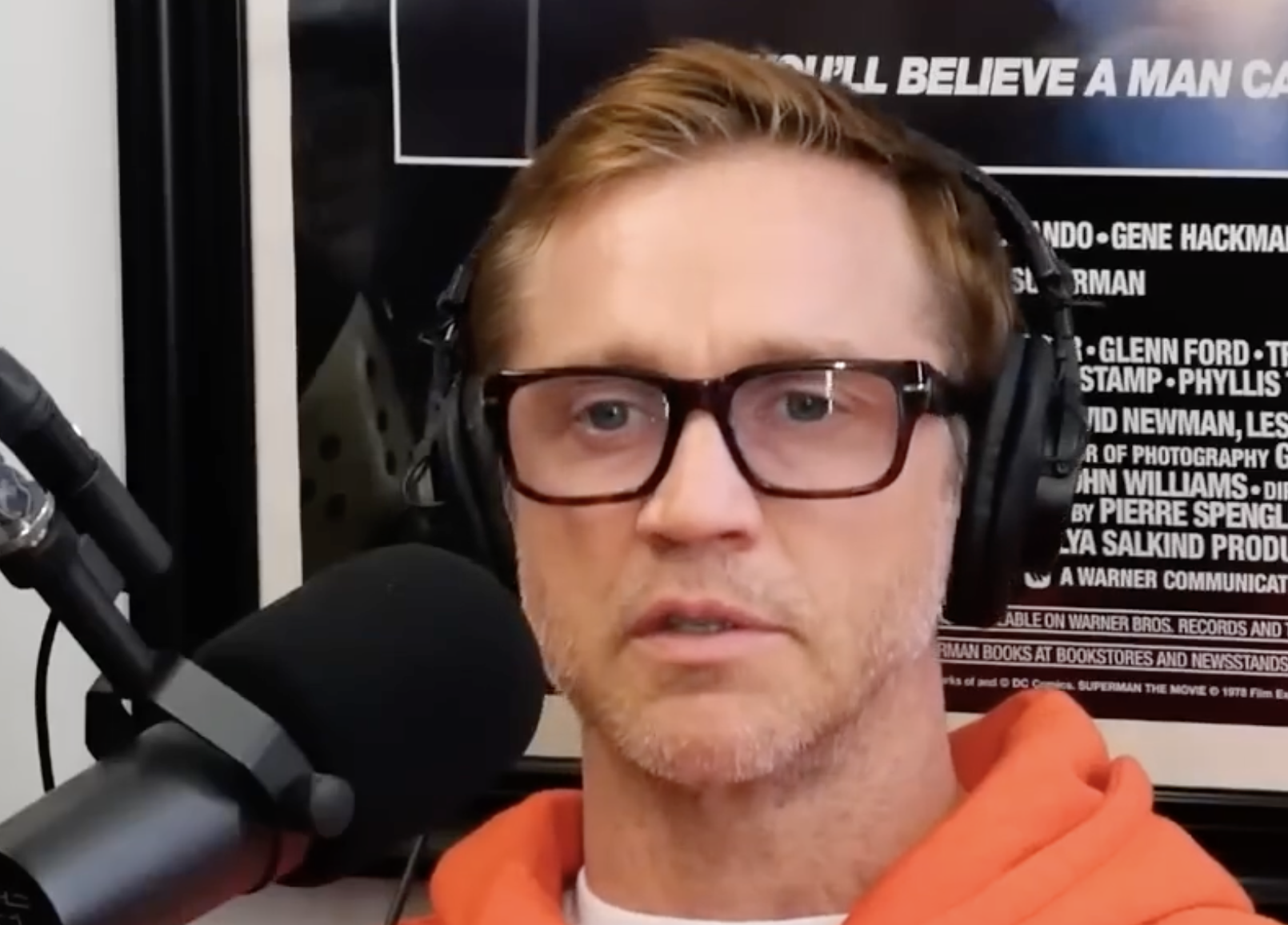 A man with reddish-brown hair wearing thick-rimmed glasses and headphones speaks into a microphone.