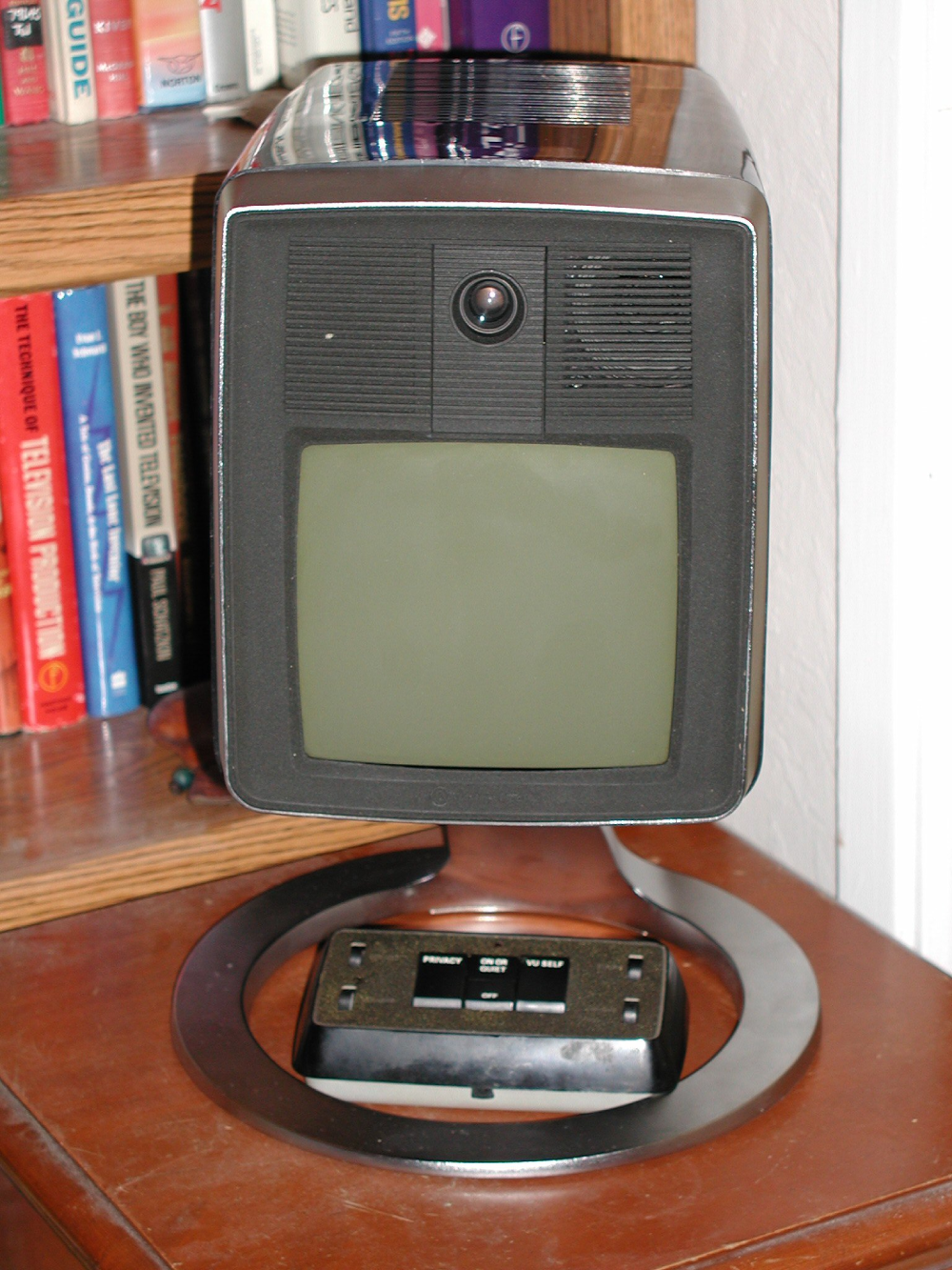 Vintage videophone on a circular stand with a built-in camera above a small monitor and a base control panel with privacy and volume settings.