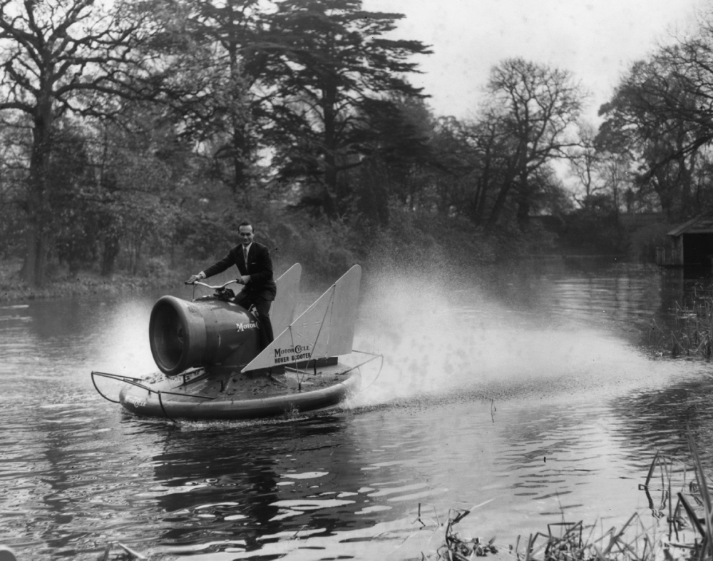 A man in a suit rides a Motor-Cycle Hover-Scooter across a river, creating a spray of water.