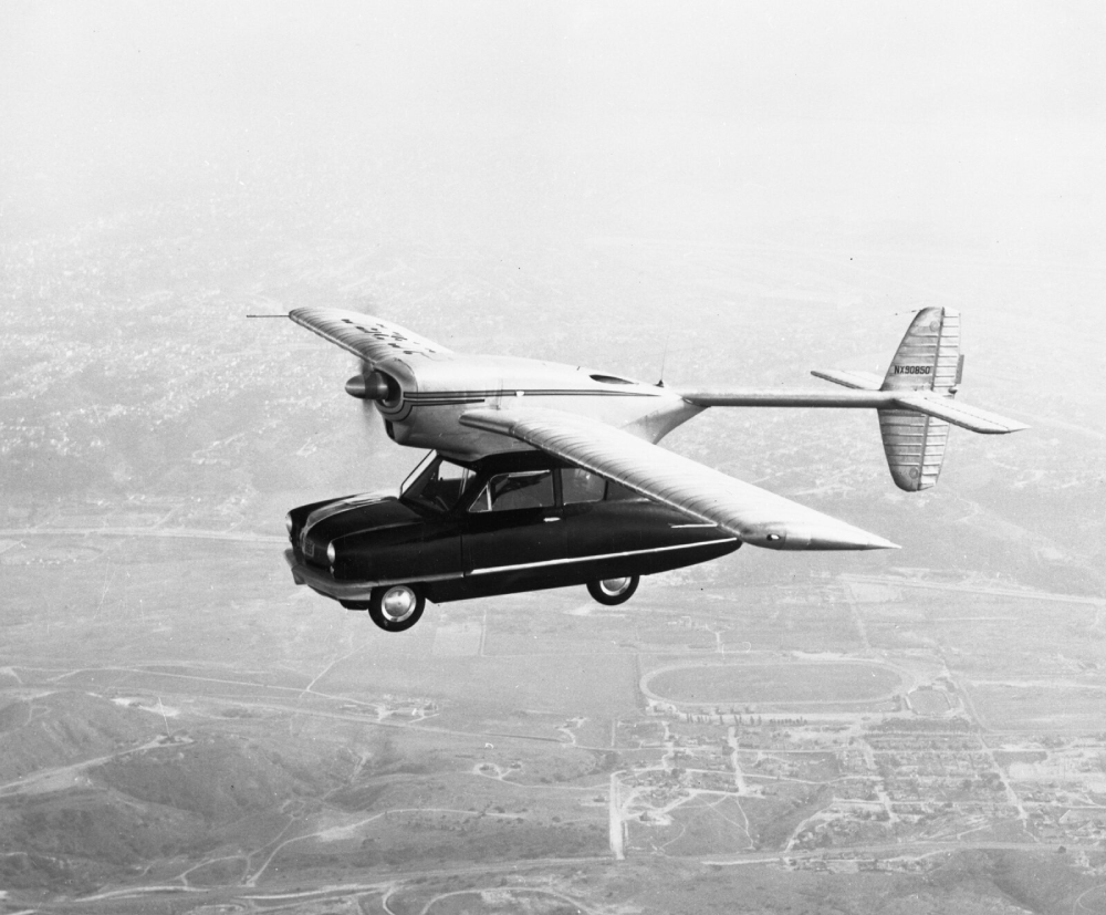A flying car, which is an automobile attached to a small airplane wing and tail assembly, in flight over a landscape.