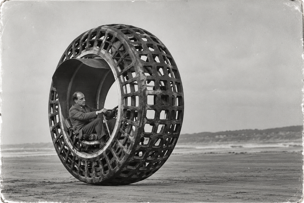 A man in a suit drives a large, circular monowheel vehicle with a grid-patterned exterior on a sandy beach.