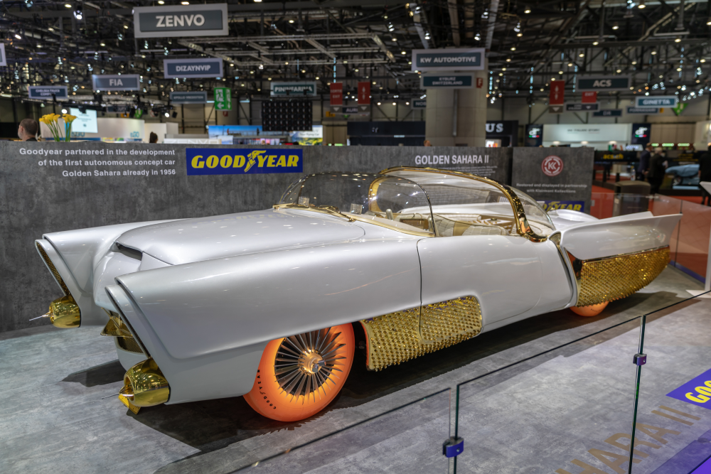 The Golden Sahara II concept car, a futuristic silver vehicle with gold accents and translucent orange tires, on display at an automotive show.