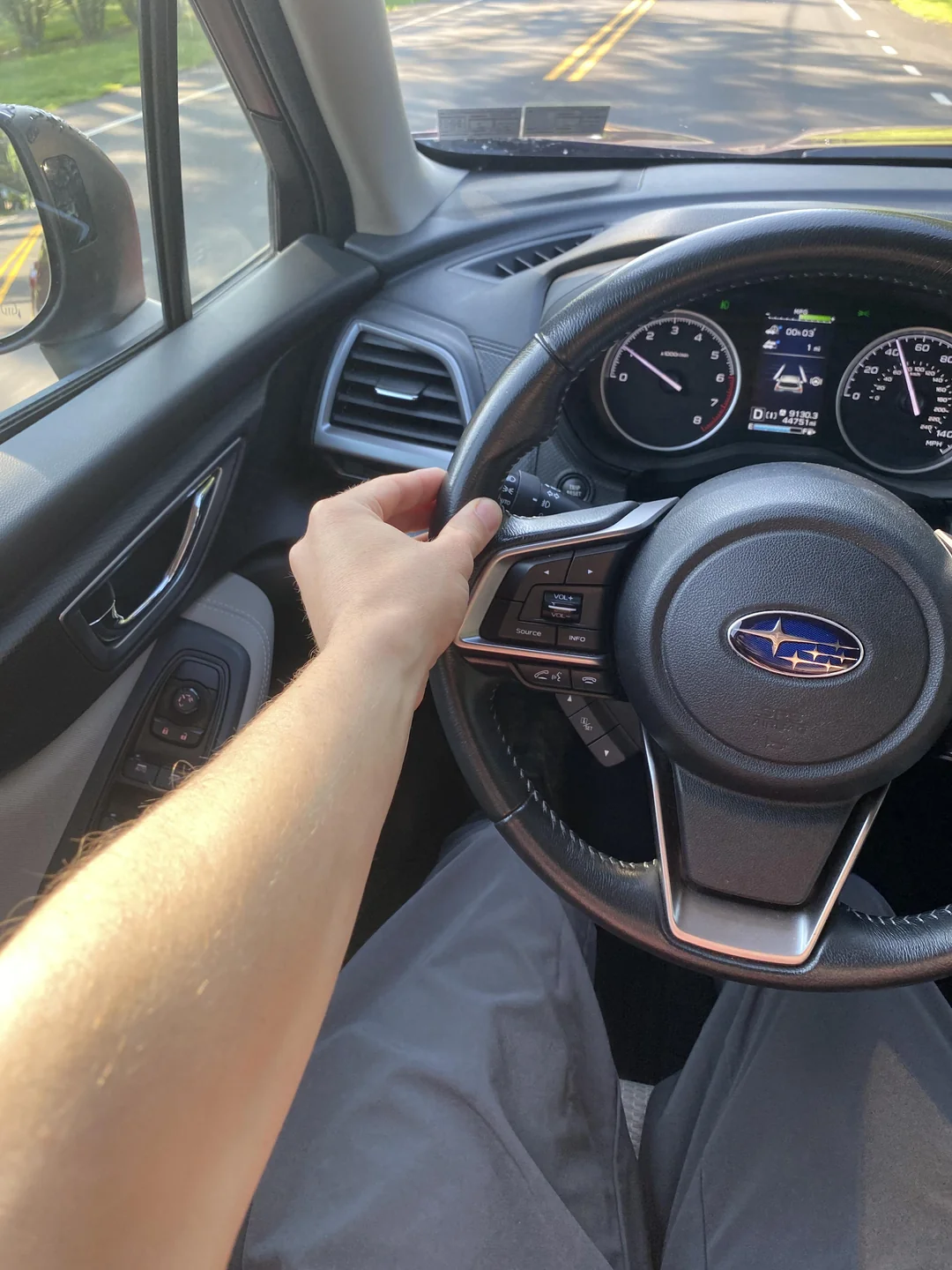 A person’s hand on the steering wheel of a Subaru car, viewed from the driver’s seat.