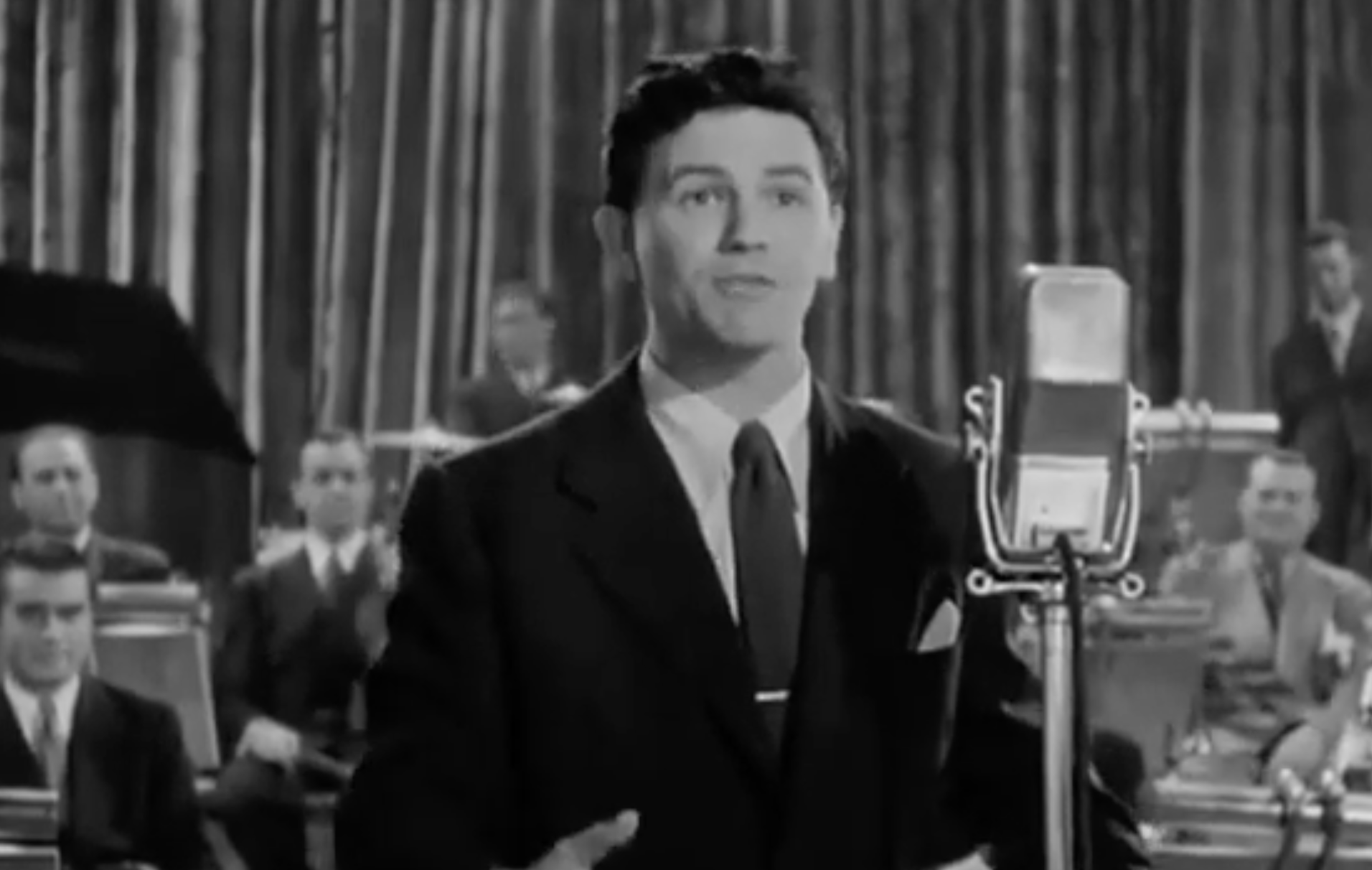A man in a suit sings into a vintage microphone in front of a big band.