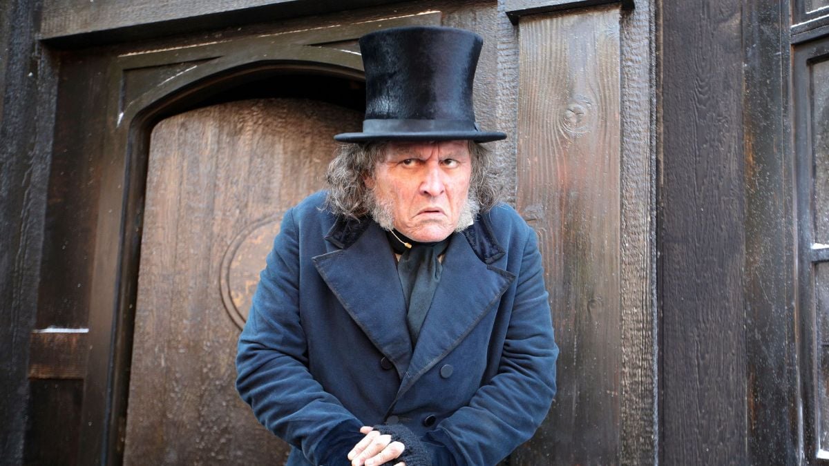 A stern man in a tall black top hat and dark coat stands hunched near a wooden door, hands clasped, with an intense, suspicious expression on his face.
