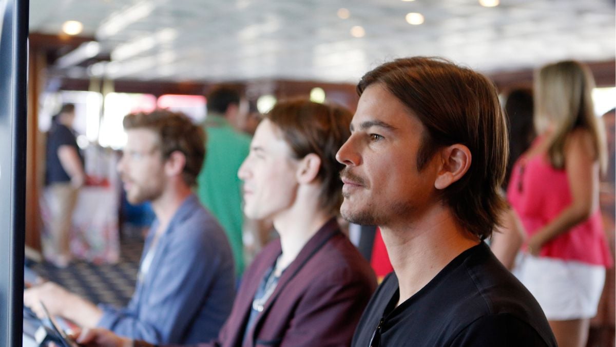 Three people sit side by side, focused on screens in front of them. The background is softly blurred, with other people standing and mingling in a well-lit indoor space.