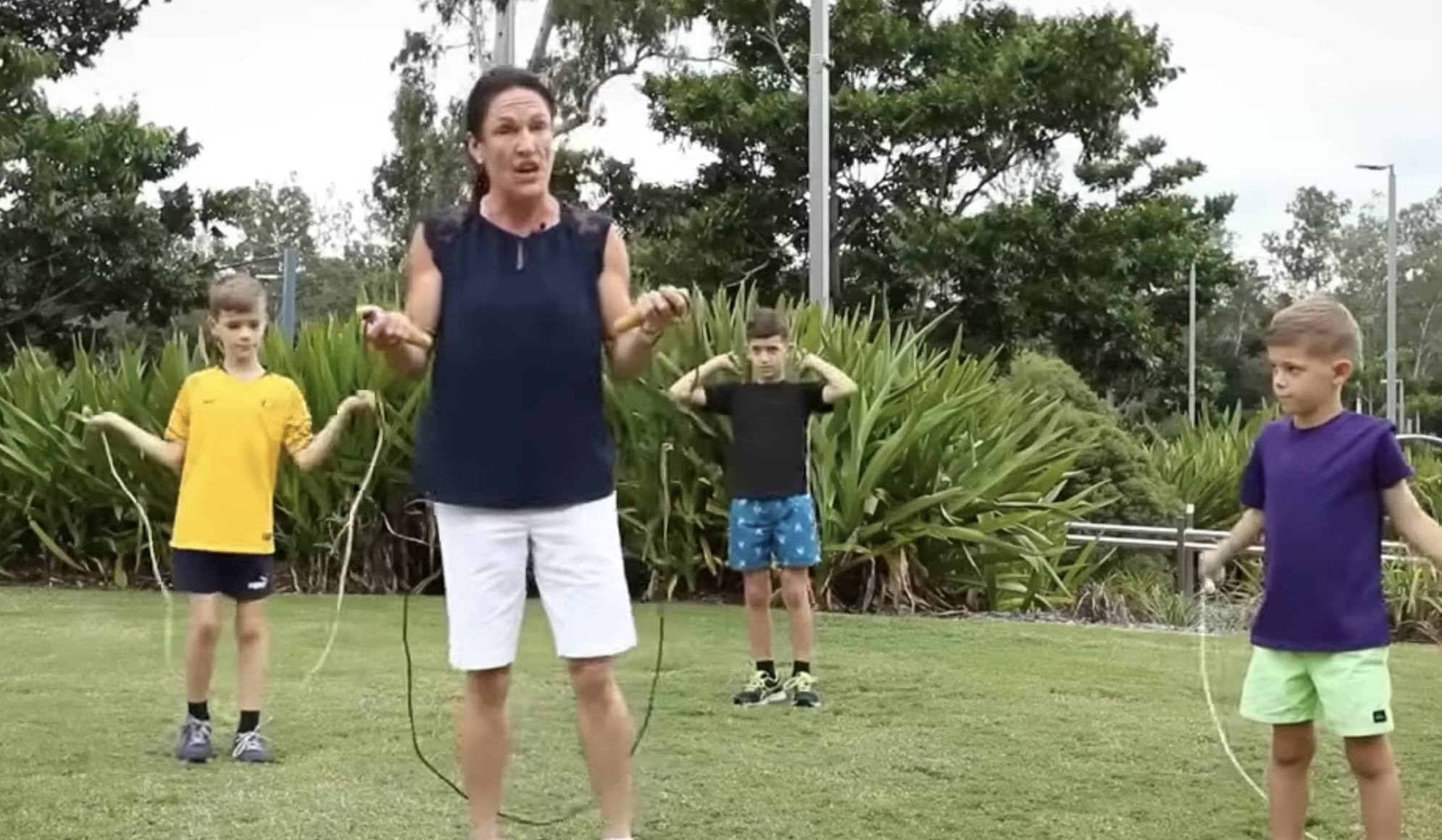 A woman in a navy top and white shorts demonstrates jump rope techniques to three young boys on a grassy field.