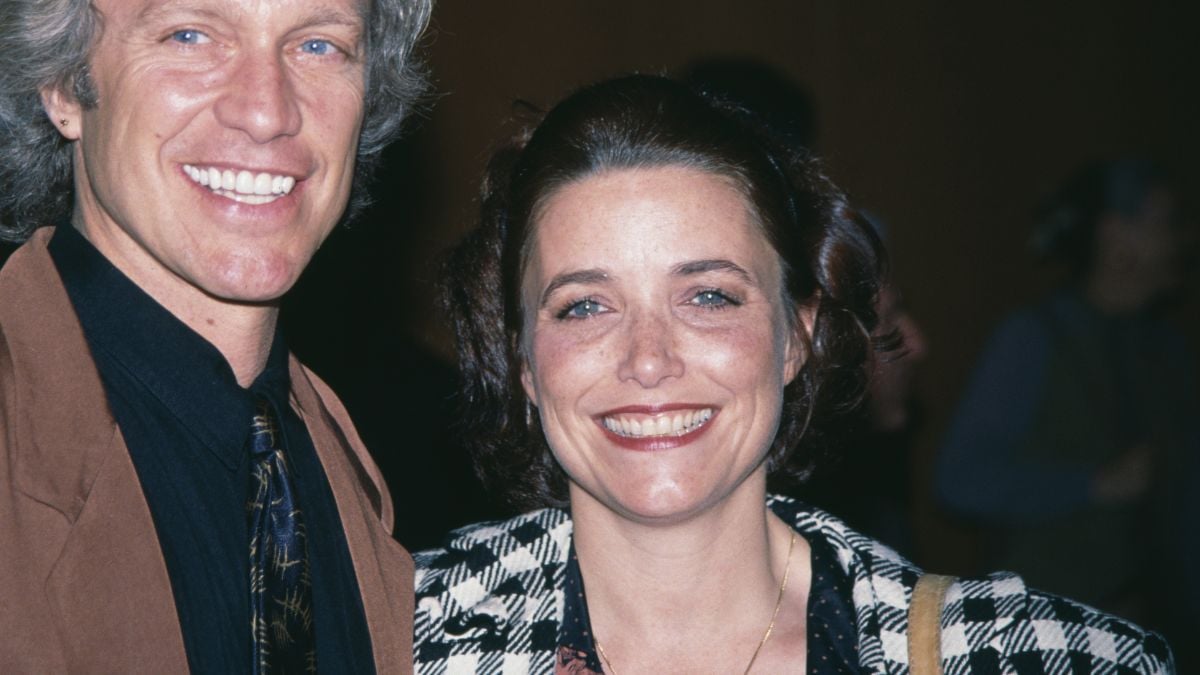 A man in a brown jacket and a woman in a black and white checkered coat smile at the camera at an indoor event.